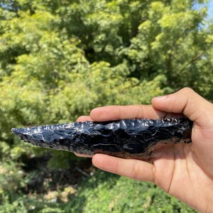 Pode incluir: Uma lâmina de obsidiana preta com uma ponta afiada e uma borda serrilhada lascada. A superfície da pedra é lisa e reflexiva, exibindo um acabamento brilhante com padrões de lascas distintos ao longo do comprimento da ferramenta.