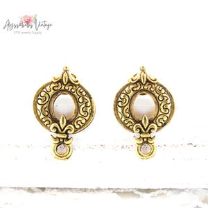 Może przedstawiać: Para złotych kolczyków z filigranowym wzorem i małym, przezroczystym kamieniem wiszącym. Kolczyki mają kształt koła z motywem lilii heraldycznej na dole.