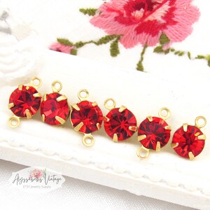 May include: Six red crystal connectors with gold-colored metal settings. Each connector features a round, faceted red crystal. The connectors have small loops on each side for attaching to other jewelry components. The background includes a white surface and a pink floral embroidery.