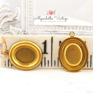 May include: Two gold-toned oval-shaped metal picture frames with a beveled edge. The frames are approximately 1 inch in diameter and have a loop at the top for hanging. The frames are shown on a ruler with the numbers 1 and 2 visible.