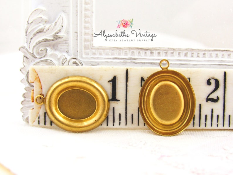 May include: Two gold-toned oval-shaped metal picture frames with a beveled edge. The frames are approximately 1 inch in diameter and have a loop at the top for hanging. The frames are shown on a ruler with the numbers 1 and 2 visible.