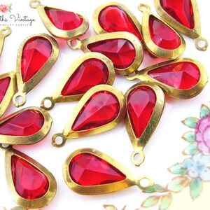 May include: A group of 15 red glass teardrop-shaped pendants with gold metal settings. The pendants are arranged on a white surface with a floral pattern.