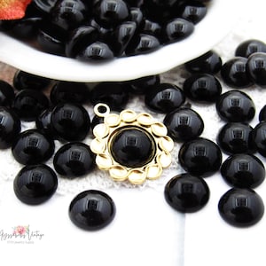 Puede incluir: Una colección de cuentas cabujón negras, lisas y redondas, esparcidas alrededor de un colgante de metal dorado con un borde festoneado y una incrustación central de cabujón negro. El texto Ayssabess Vintage Etsy Jewelry Supply es visible abajo.