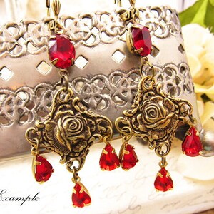 May include: Gold tone earrings with red glass teardrop and rose accents. The earrings feature a dangling rose design with multiple red teardrop crystals.
