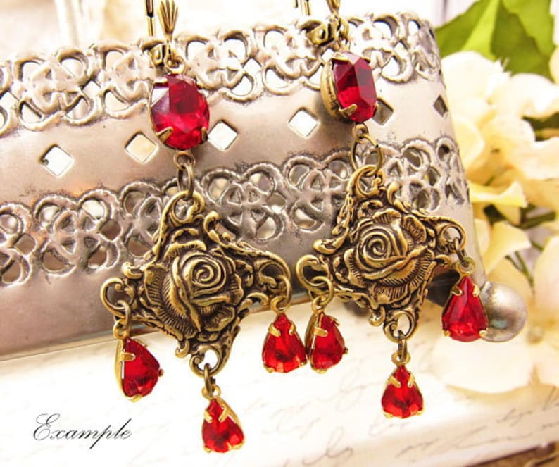 May include: Gold tone earrings with red glass teardrop and rose accents. The earrings feature a dangling rose design with multiple red teardrop crystals.