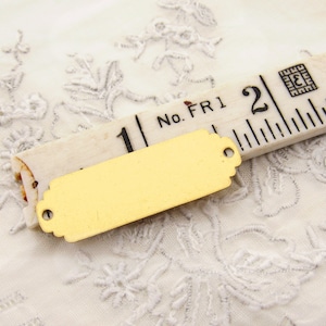 May include: A gold-coloured metal tag with a rectangular shape and rounded edges. The tag has two holes for attaching it to something. The tag is lying on a white fabric with a floral pattern. The tag is next to a wooden ruler with the numbers "1" and "2" visible.