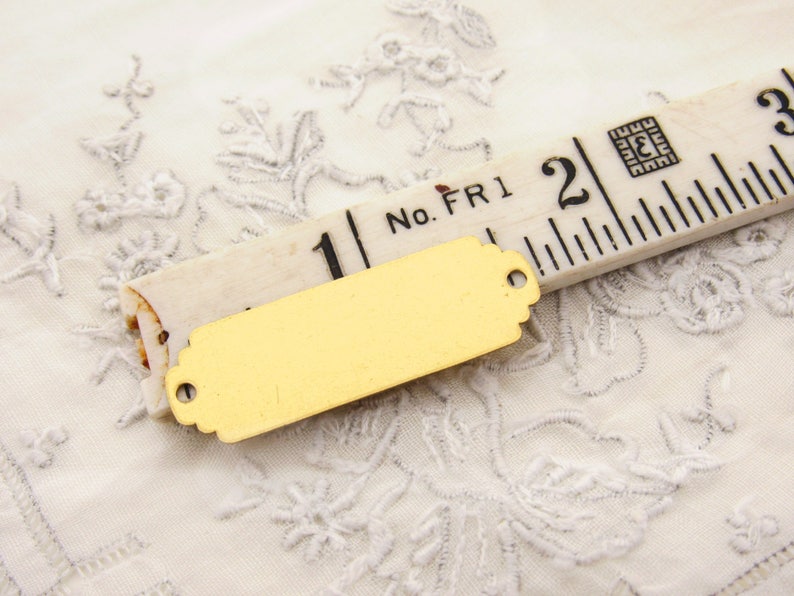 May include: A gold-coloured metal tag with a rectangular shape and rounded edges. The tag has two holes for attaching it to something. The tag is lying on a white fabric with a floral pattern. The tag is next to a wooden ruler with the numbers "1" and "2" visible.