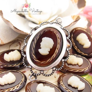 May include: A collection of vintage cameo pendants and cabochons. The oval-shaped cameos feature a white profile against a dark brown background, set in silver-toned frames. The cabochons have a similar design. The Etsy shop name "Alyssabeths Vintage" is visible.