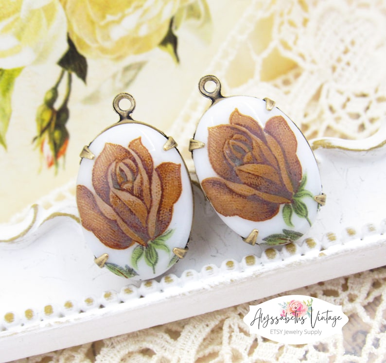May include: Two oval-shaped porcelain charms with a brown rose design and gold accents. The charms have a hole at the top for hanging.