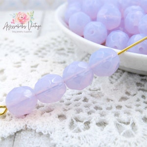 May include: A strand of light purple faceted glass beads on a white crocheted surface. The beads are strung on a gold wire.