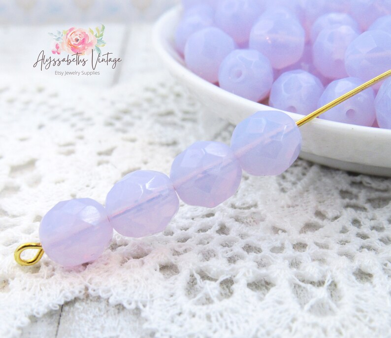 May include: A strand of light purple faceted glass beads on a white crocheted surface. The beads are strung on a gold wire.