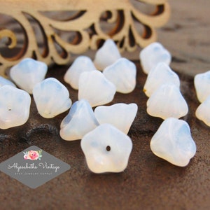 May include: A collection of 15 white opaque glass flower shaped beads. The beads have a slightly iridescent finish and a hole in the center for stringing.