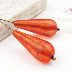 May include: Close-up of two orange teardrop-shaped beads with a crackle effect, attached to gold-colored wires. The beads have a small, round, gold-colored bead at the bottom. The background is white with a textured surface.