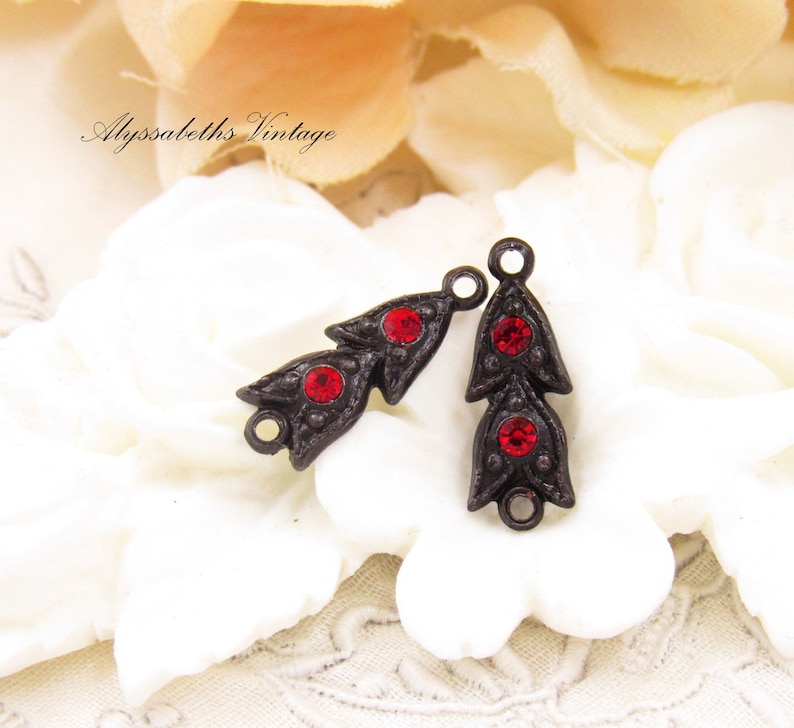 Puede incluir: Dos abalorios de metal de estilo antiguo con detalles de strass rojos. Cada abalorio presenta un dise&ntilde;o de hoja estilizado con tres strass rojos.