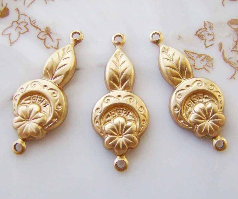 May include: Three gold-toned metal charms with a floral design and a leaf detail. Each charm has a loop at the top and bottom for attaching to jewelry.
