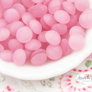 Op de afbeelding: Een verzameling kleine, ronde, doorschijnende roze glazen cabochons met een glad, gewelfd oppervlak en subtiele horizontale strepen. Deze ronde kralen liggen op een hoop in een witte schaal en hebben een consistente lichtroze kleur.