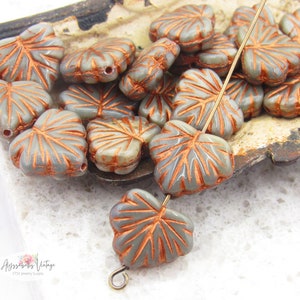 May include: A close-up of a pile of gray glass leaf-shaped beads with copper accents. The beads are arranged on a metal tray with a gold wire running through them.