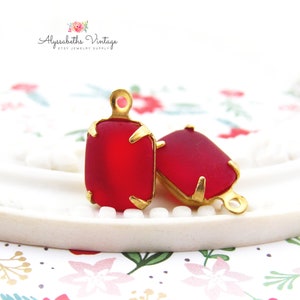 May include: Two red glass cabochons with gold settings and jump rings. The cabochons are rectangular and have a slightly frosted finish.