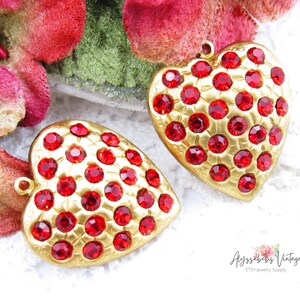 May include: Two gold heart-shaped pendants with red rhinestone accents. The pendants are on a white background.