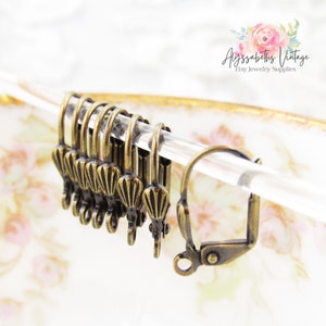 Może przedstawiać: Zbliżenie dziesięciu antycznych haczyków do kolczyków wykonanych z mosiądzu z ozdobnym wzorem muszli. Haczyki są prezentowane na przezroczystej szynie.