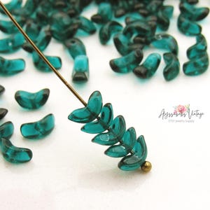 May include: Teal-colored glass beads in a crescent moon shape. A few beads are strung on a gold-colored wire, showcasing their curved design. The beads are scattered on a white surface, with the Etsy shop name "Alyssabeths Vintage" in the background.