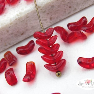 May include: Close-up of red, crescent-shaped beads. Some beads are strung on a gold-colored pin, while others are scattered on a white surface. The beads are translucent and have a glossy finish, perfect for jewelry making.