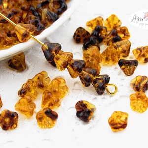 May include: Close-up of amber-colored glass beads with a flower-like shape. Some beads are strung on a gold-colored wire, while others are scattered on a white surface. The beads have a translucent quality.
