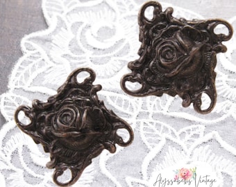May include: Two antique bronze metal rose flower embellishments with intricate details. Each embellishment has a rose in the center and a decorative frame. The embellishments have four holes for attaching to a project.