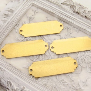 May include: Four gold-coloured metal rectangular tags with rounded corners and two holes on each tag. The tags are on a white surface with a decorative border.
