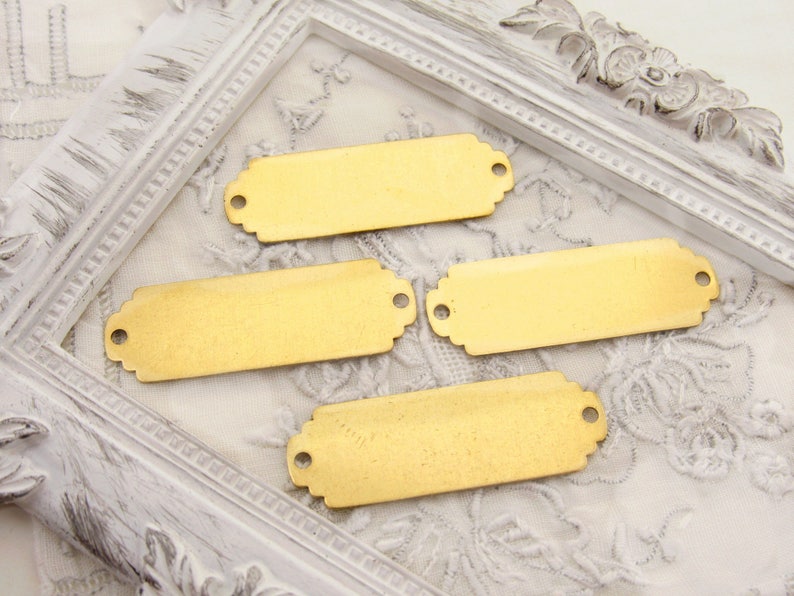 May include: Four gold-coloured metal rectangular tags with rounded corners and two holes on each tag. The tags are on a white surface with a decorative border.