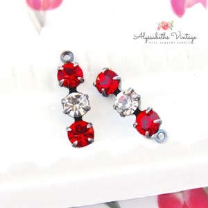 Peut inclure: Deux attaches de boucles d'oreilles argentées avec trois strass rouges et transparents chacune. Les strass sont disposés verticalement avec un strass rouge en haut et en bas et un strass transparent au milieu.