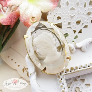 May include: Oval-shaped cameo pendant with a clear, faceted glass frame and gold-colored metal accents. The cameo features a white profile of a woman. The pendant is a jewelry supply item.