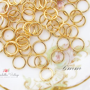 May include: A collection of gold-colored, 6mm jump rings, perfect for jewelry making. The rings are open and ready to be connected. The image is set on a white and gold surface. The text "6mm" is visible.