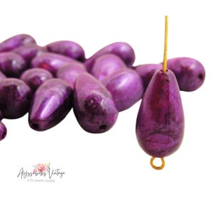 May include: A close-up of a pile of purple pearlescent teardrop-shaped beads. One bead is hanging from a gold wire loop.