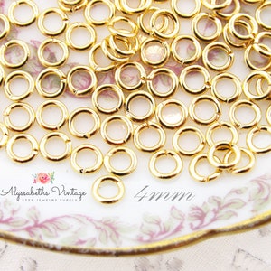May include: A close-up of a pile of gold jump rings, 4mm in diameter, on a white and pink floral patterned plate. The jump rings are a popular jewelry making supply.