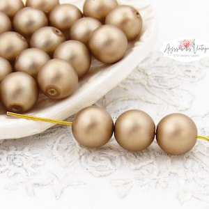 May include: A collection of golden-colored, round beads, some displayed in a white, scalloped dish. Three beads are strung on a gold-colored wire, showcasing their smooth, matte finish. The background is a white, lace-like fabric.