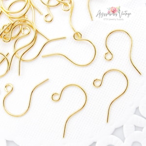 May include: Gold-toned earring hooks, also known as ear wires, are shown on a white surface with a lace pattern. The hooks are made of metal and have a simple, classic design.