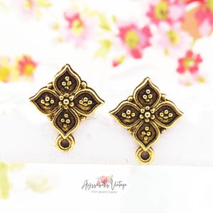 Może przedstawiać: Dwa antyczne, złote elementy kolczyków w kształcie kwiatów. Każdy element ma czteropłatkowy wzór z małymi okrągłymi detalami i pętlą na dole. Tło jest rozmyte z różowymi i białymi kwiatami. Nazwa sklepu Etsy "Alyssabeths Vintage" znajduje się na dole.