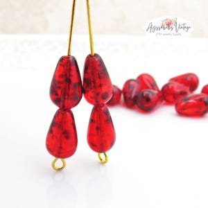 Op de afbeelding: Close-up van rode, druppelvormige kralen met donkere spikkels, geregen op goudkleurig draad. De kralen zijn gegroepeerd, met een paar verspreid op de achtergrond. Het Etsy Jewelry Supply-logo is rechtsboven zichtbaar.