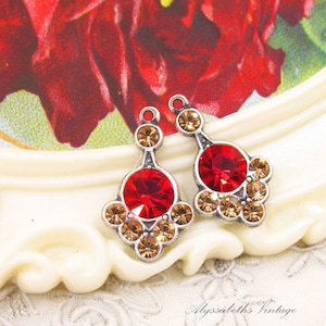 May include: Two silver-toned metal jewelry charms featuring a large, round, faceted red crystal at the center, surrounded by smaller, round, faceted champagne-colored crystals. Each charm has a small loop at the top for attachment purposes.