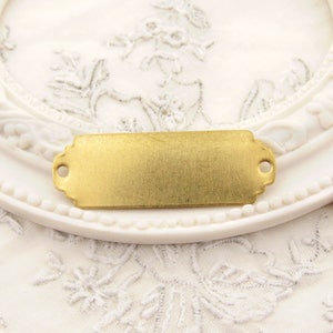 May include: A gold-toned metal tag with rounded corners and two small holes at the top. The tag is blank and ready for personalisation.