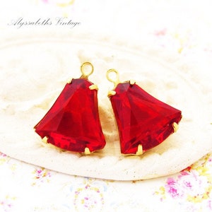 May include: Two faceted, trapezoidal red glass pendants set in gold-toned metal prongs. Each pendant features a small circular loop at the top for attachment. The vibrant red stones have a geometric cut that reflects light across their surfaces.