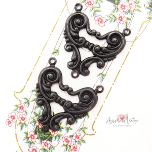 May include: Two black metal heart-shaped embellishments with intricate scrollwork and three jump rings. The embellishments are on a floral patterned surface.