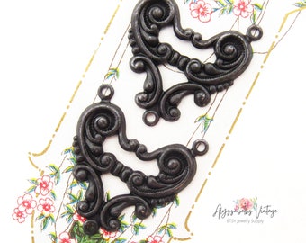 May include: Two black metal heart-shaped embellishments with intricate scrollwork and three jump rings. The embellishments are on a floral patterned surface.