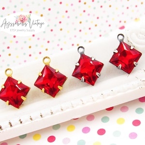 May include: Four red square-shaped glass charms with gold and silver settings. The charms have a loop at the top for attaching to jewelry.