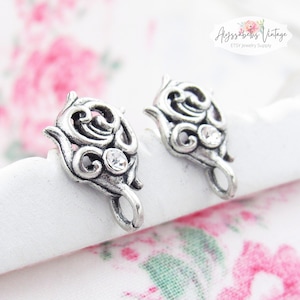 May include: Two silver-toned metal earring findings with a decorative floral design and a clear rhinestone accent.  The findings have a loop at the bottom for attaching to earrings.