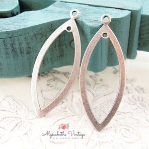 May include: Two silver-toned metal earring blanks in the shape of an oval with a pointed tip. The blanks have a hole at the top and a smaller hole in the center.