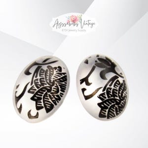 May include: Pair of oval-shaped, white glass cabochons with black floral designs. The cabochons are approximately 1 inch in diameter.
