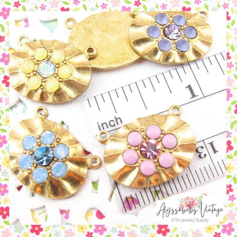 May include: Four gold-toned oval-shaped metal charms with a scalloped edge. Each charm features a flower design with a central rhinestone and surrounding colored stones. The charms are approximately 1 inch in length and have two jump rings for attaching to jewelry.
