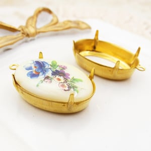 May include: Two oval-shaped gold-tone metal jewelry findings. One finding has a cream-colored oval cabochon with a floral design in blue, purple, and pink. The other is an empty gold-tone metal setting.